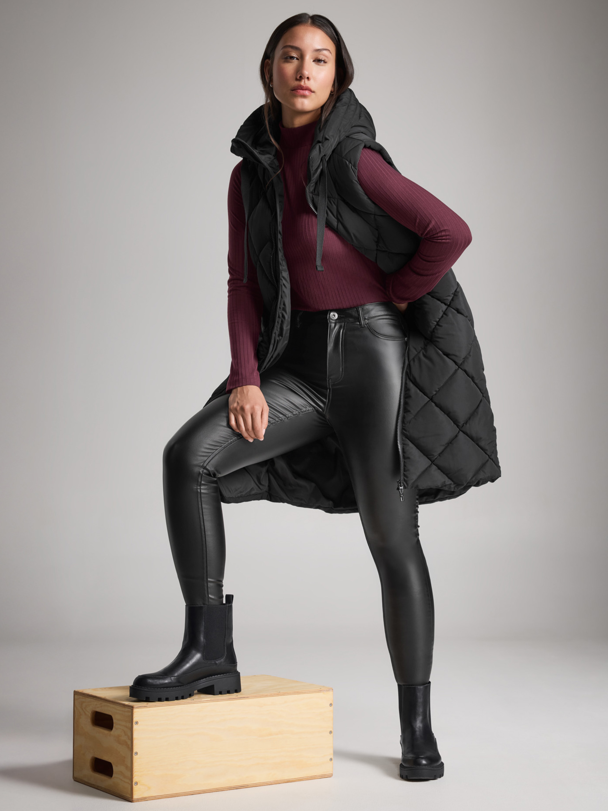 Femme en gilet matelassé noir, jean en cuir et bottes, un pied sur une boîte.