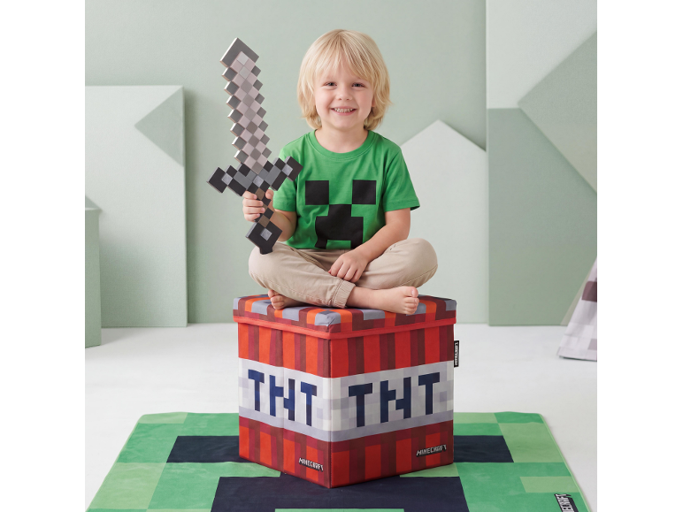 Enfant souriant avec épée jouet, assis sur une boîte de rangement en forme de bloc TNT, sur un tapis à thème jeu vidéo.