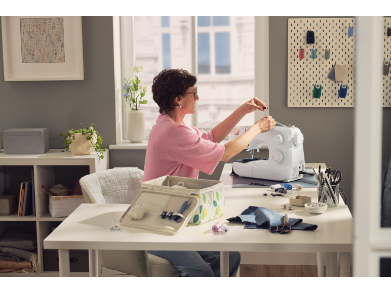 Femme cousant sur une machine à coudre, entourée d'accessoires de couture et de boîtes de rangement.