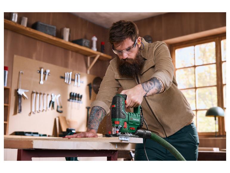 Un homme barbu utilise une scie sauteuse pour couper une planche de bois dans un atelier.