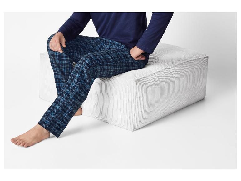 Homme en pyjama à carreaux bleus et chemise bleu marine assis sur un pouf blanc.