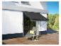 Un store banne noir, une chaise longue et un pot de fleurs sur une terrasse en bois.