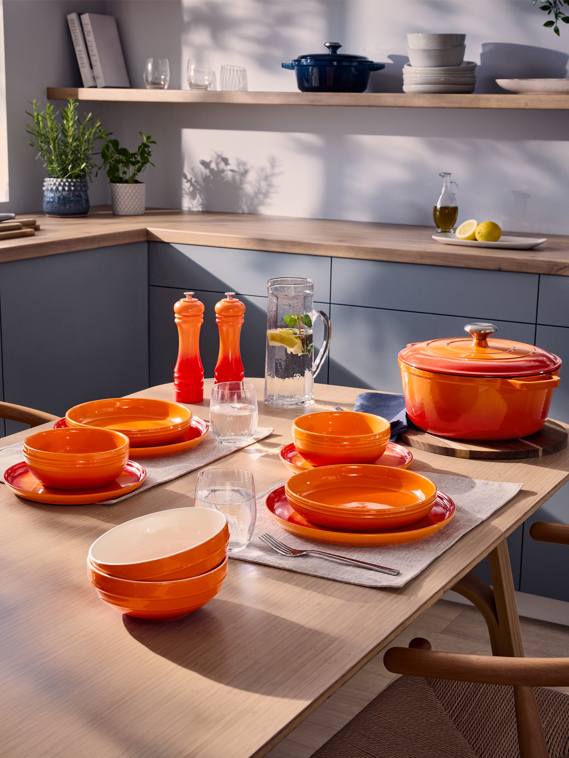 Vaisselle orange et cocotte en fonte sur une table de cuisine avec herbes et ustensiles.