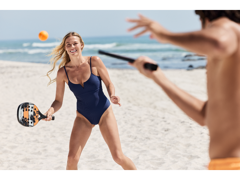 Femme en maillot de bain jouant au beach tennis avec raquette et balle sur la plage.