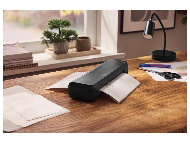 Plastifieuse noire sur une table en bois avec des papiers, des ciseaux et une lampe de bureau.