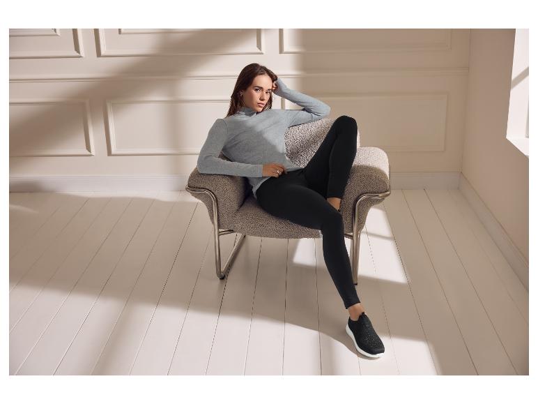 Femme en col roulé gris et leggings noirs assise dans un fauteuil.