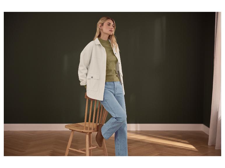 Femme en pull vert, veste blanche et jean bleu, appuyée sur une chaise en bois.