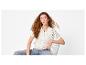 Femme rousse, portant un cardigan blanc en crochet et un jean, assise sur une chaise.