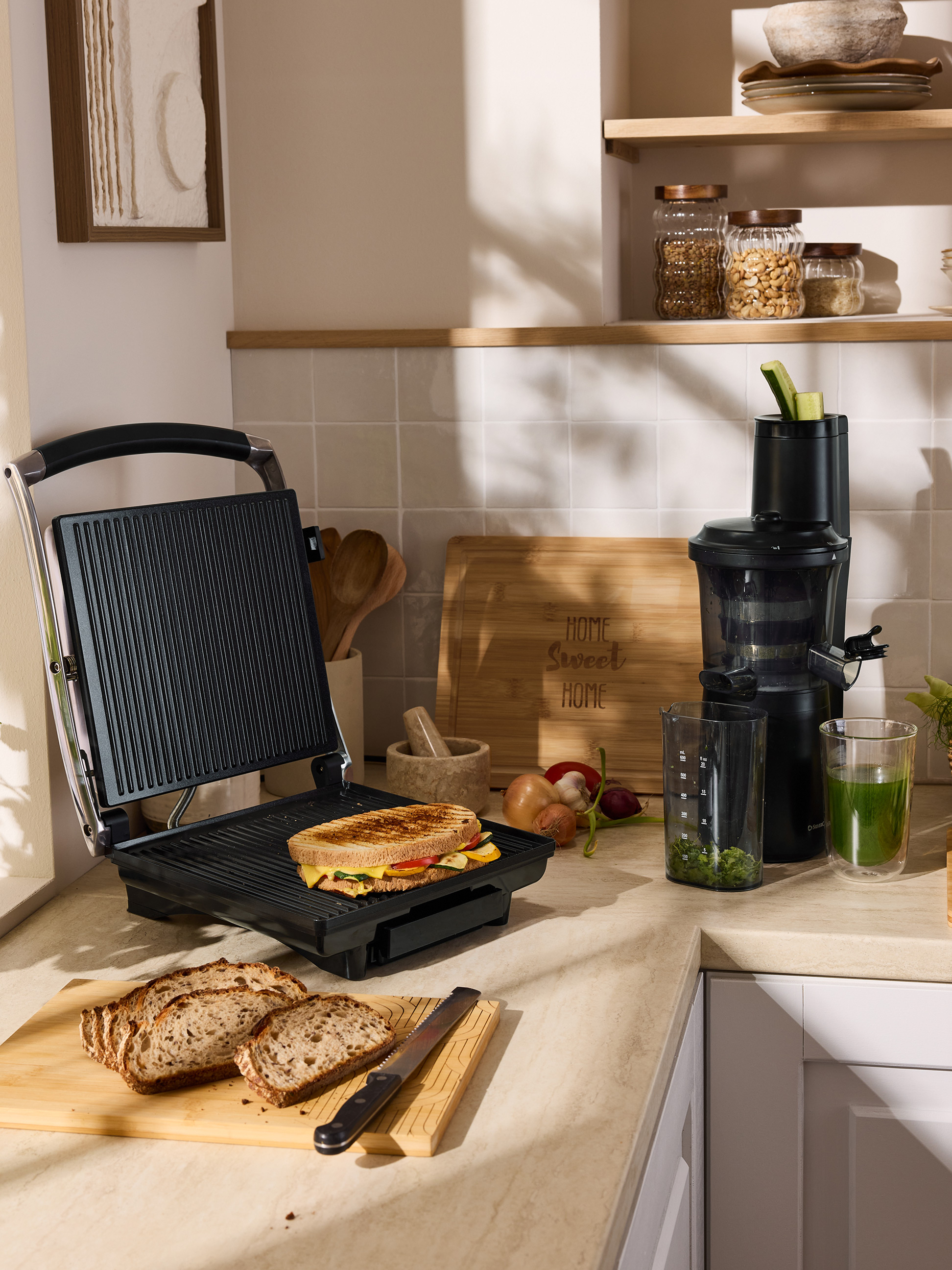 Un sandwich grillé sur un appareil à sandwichs, du pain tranché et un extracteur de jus sur un comptoir de cuisine.