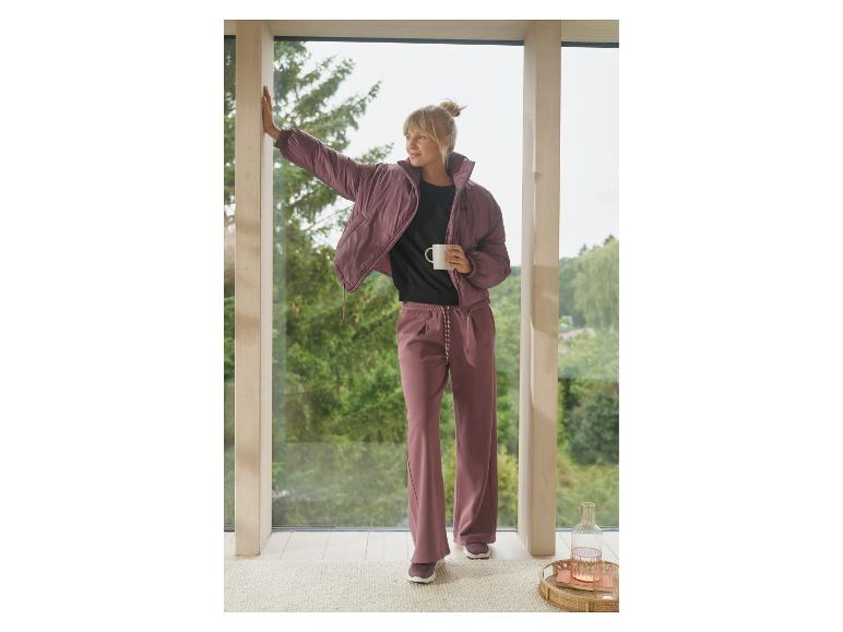 Femme en veste matelassée rose et pantalon de survêtement tenant une tasse près de la fenêtre.