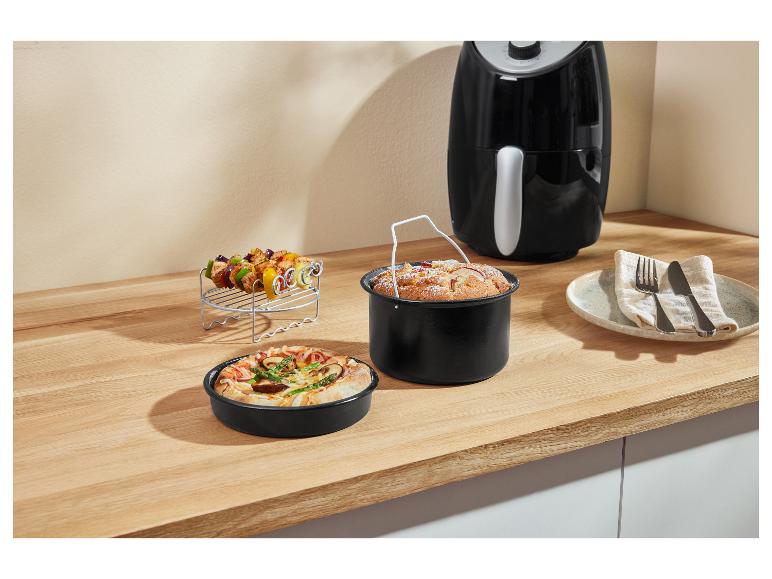 Friteuse à air avec accessoires : brochettes, moule à gâteau et plat à pizza sur un comptoir.