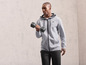 Un homme en sweat à capuche gris zippé et pantalon noir soulève une haltère.