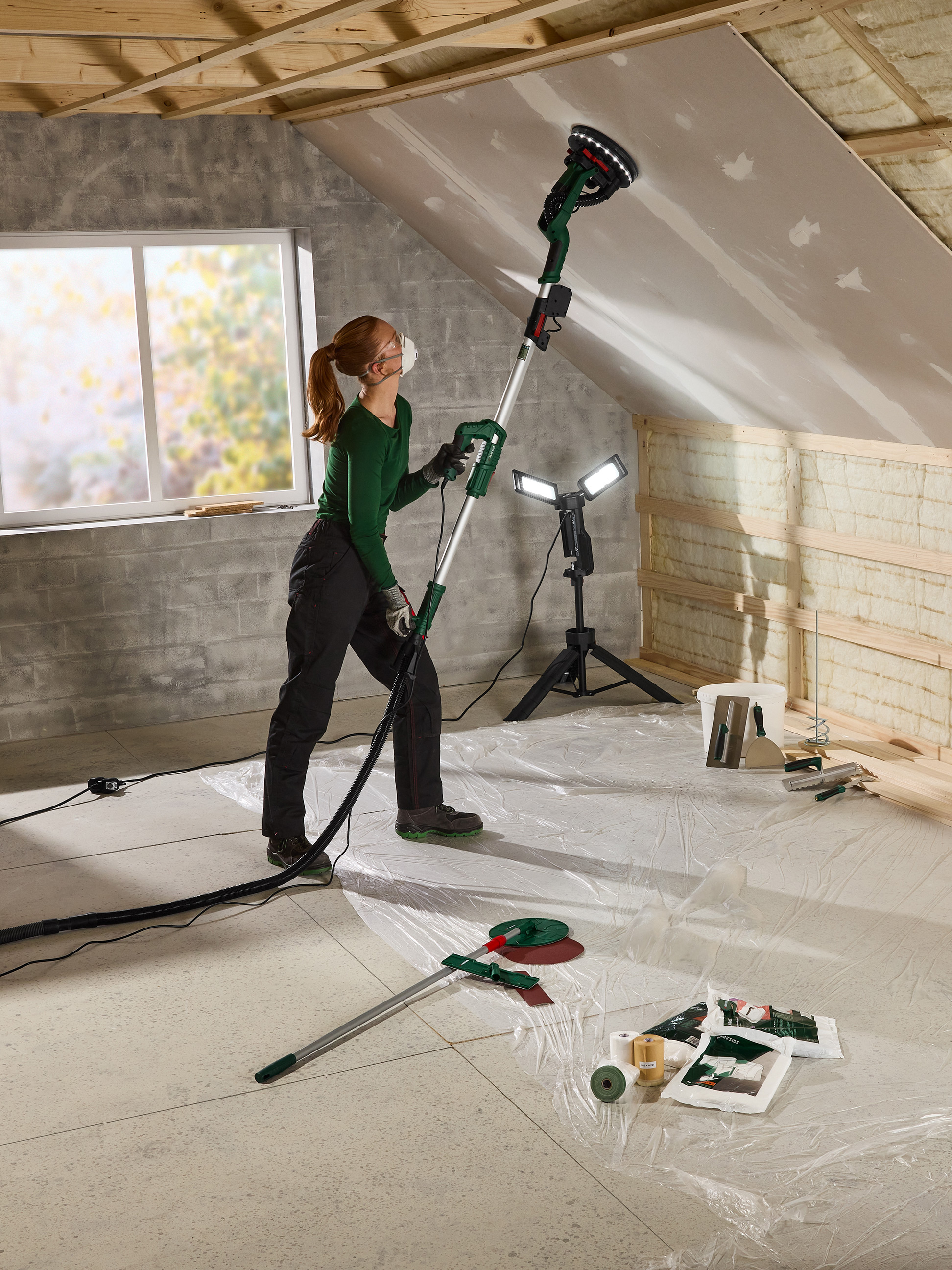 Femme ponçant un plafond de grenier avec une ponceuse à plâtre et une lampe de travail.
