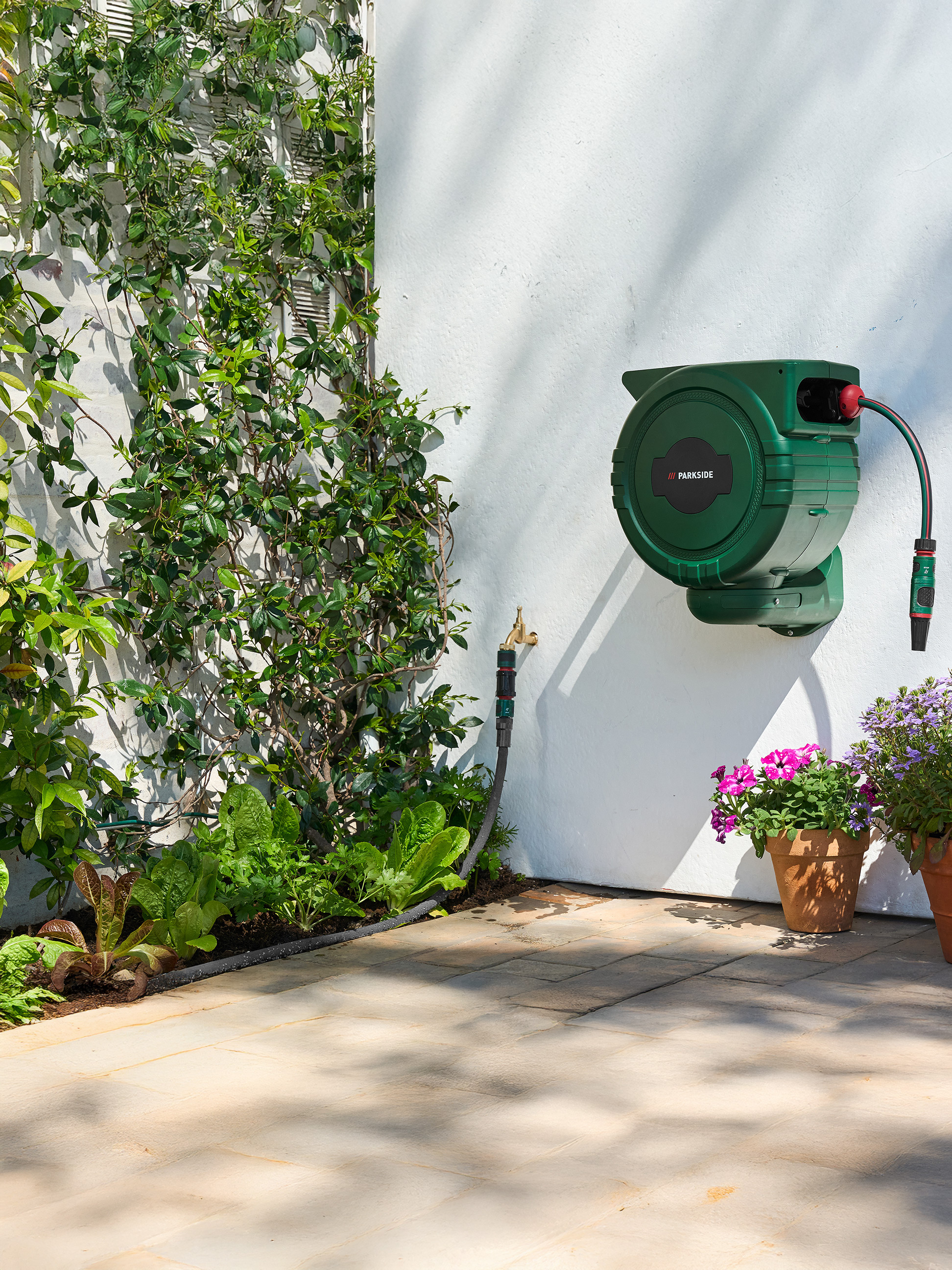 Dévidoir de tuyau d'arrosage vert PARKSIDE sur un mur blanc avec plantes grimpantes et potager.