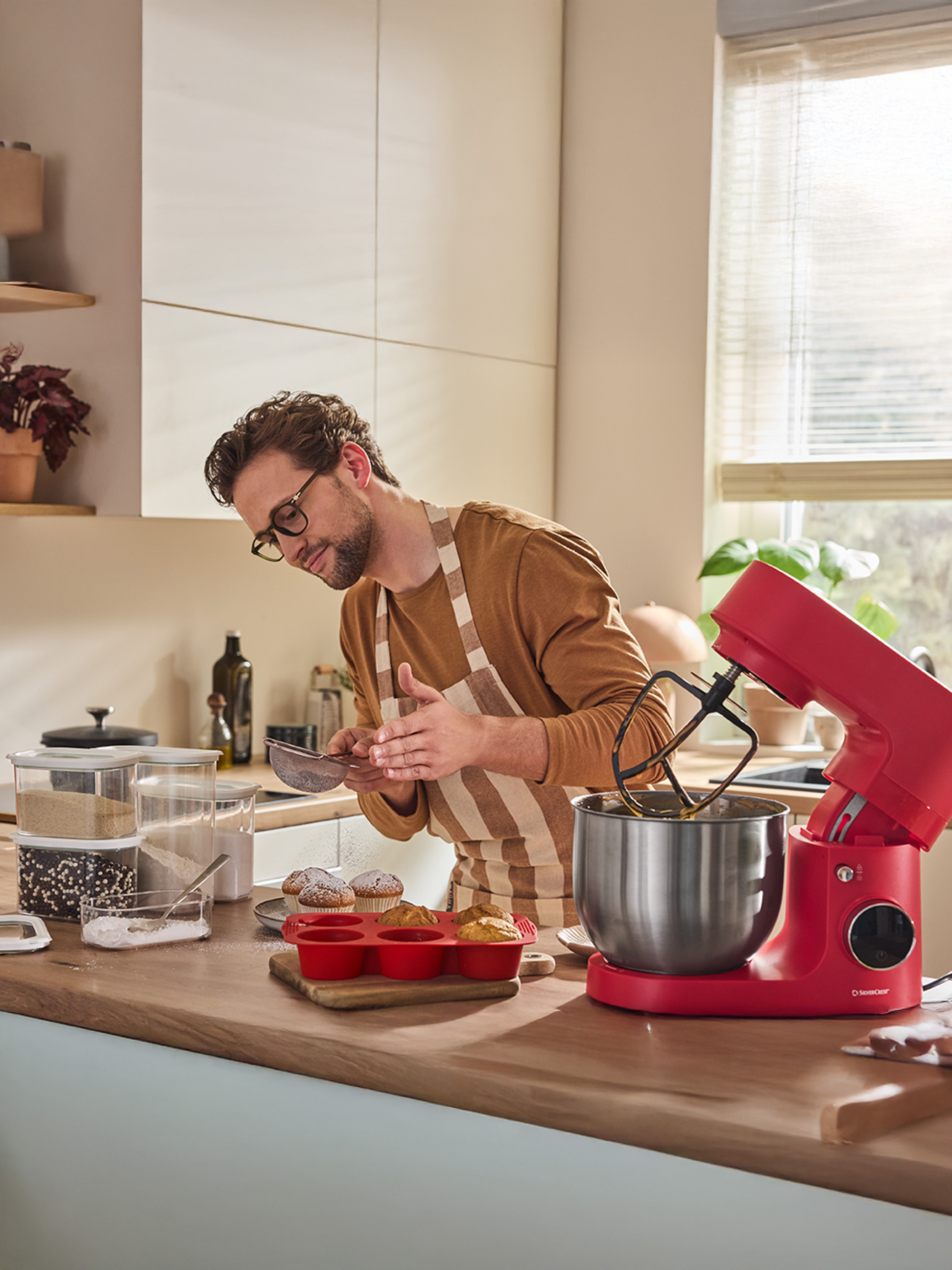 Homme saupoudre du sucre glace sur des muffins à côté d'un robot pâtissier rouge SilverCrest.