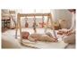 Bébé allongé sous un portique d'éveil en bois avec des jouets suspendus, une personne à côté.