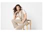 Femme en gilet et pantalon beiges, assise sur un tabouret en bois.
