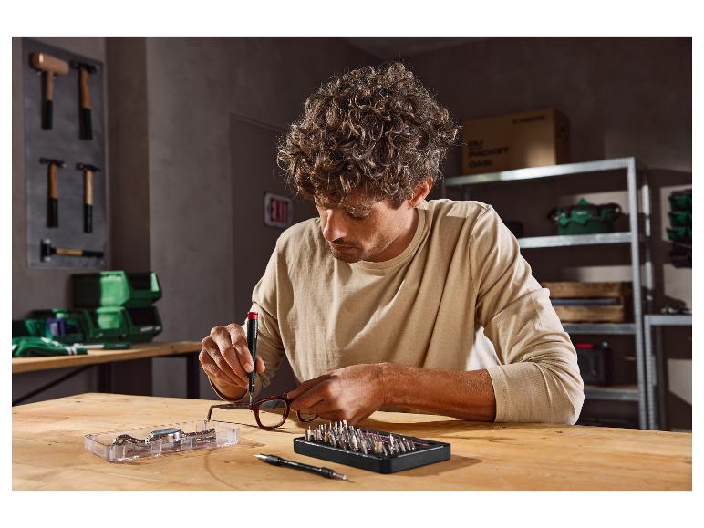Homme réparant des lunettes avec un tournevis de précision et un jeu d'embouts sur un établi en bois.