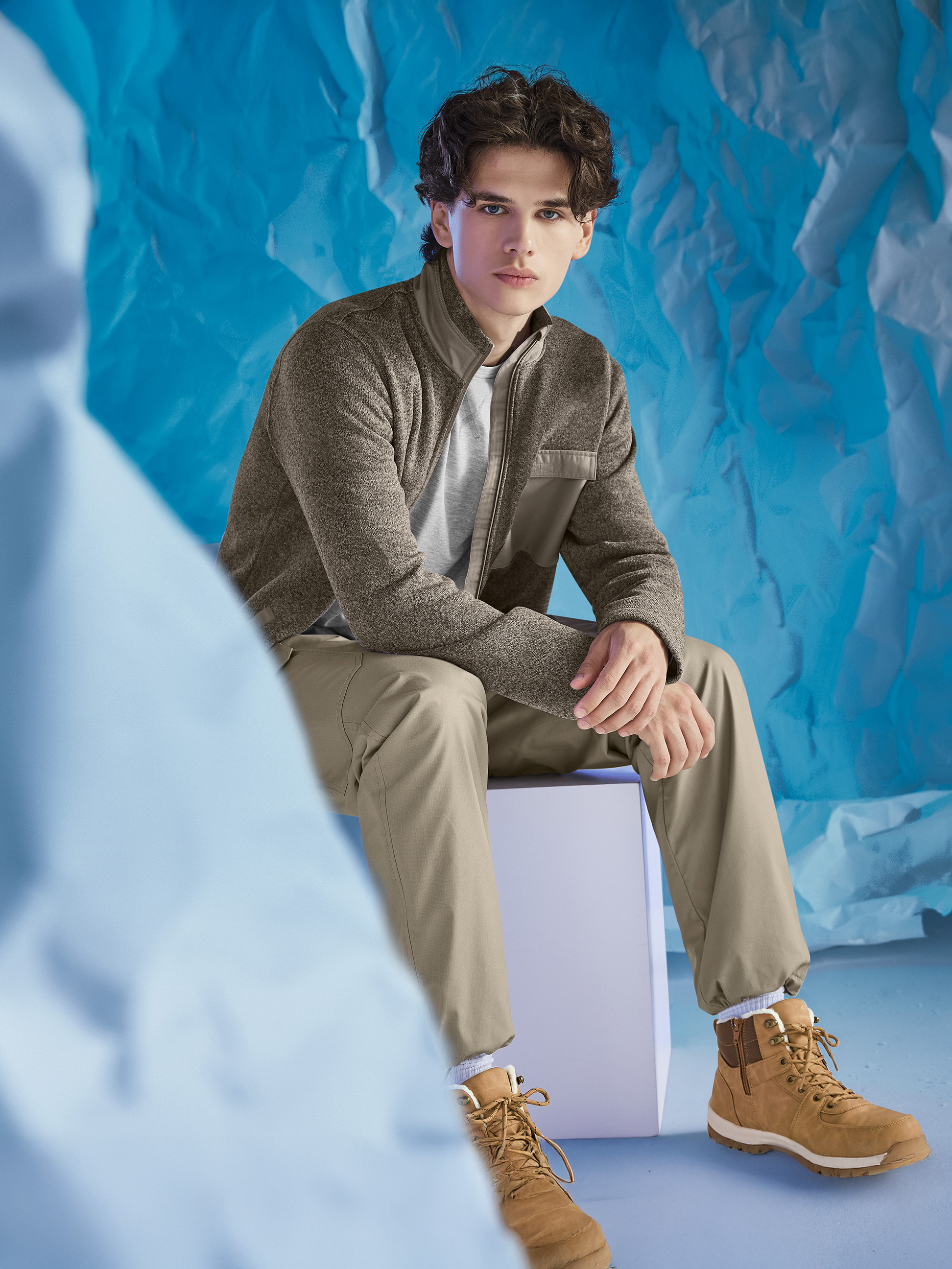 Un homme porte une veste marron, un t-shirt gris et un pantalon kaki, assis sur fond bleu.