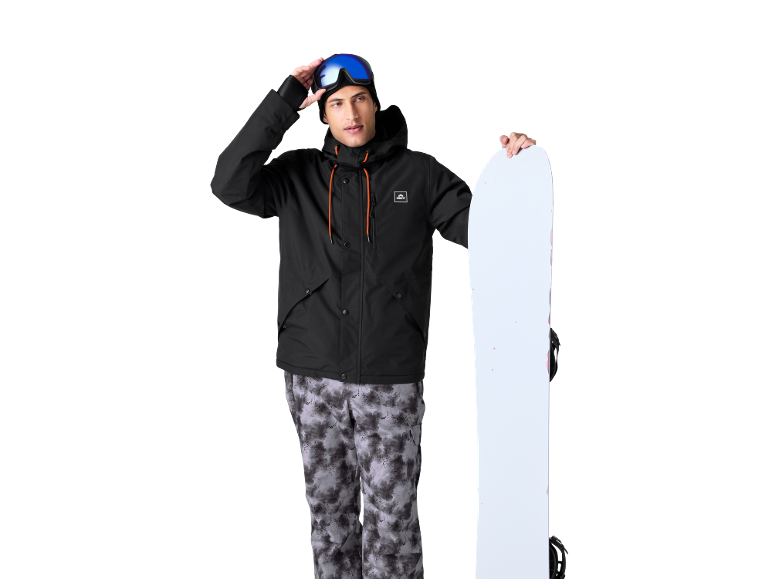 Homme en veste de ski noire et pantalon de ski à motifs tenant un snowboard.