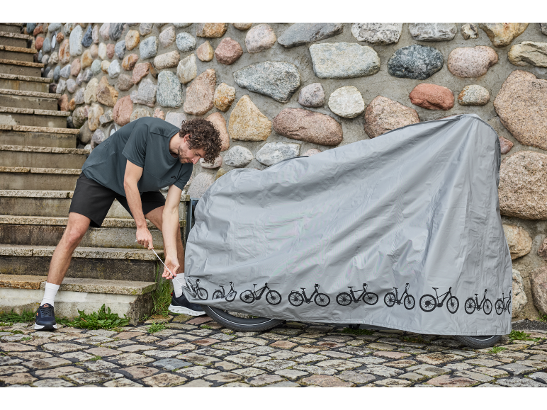 Un homme ajuste une housse de protection grise sur un vélo à côté d'un escalier en pierre.