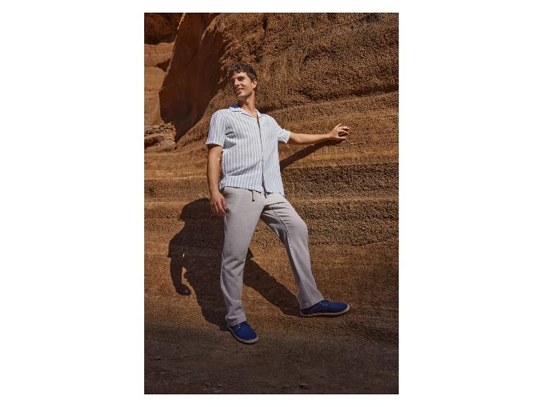 Homme en chemise rayée et pantalon gris posant contre une paroi rocheuse.