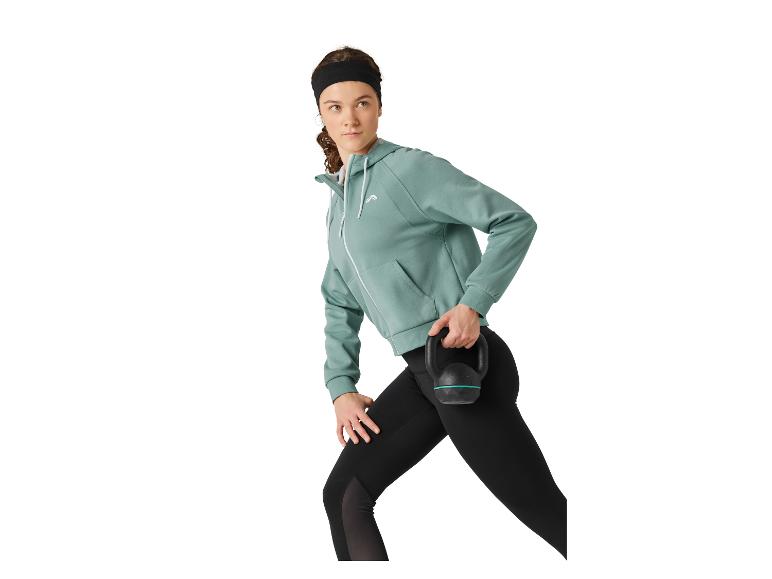Femme en sweat à capuche sarcelle et leggings noirs tenant un kettlebell, prête à s'entraîner.