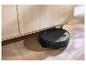 Robot aspirateur noir nettoyant sous une armoire en bois clair sur un parquet.