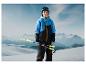 Un garçon en veste bleue et pantalon noir tient des bâtons de ski dans un paysage enneigé.