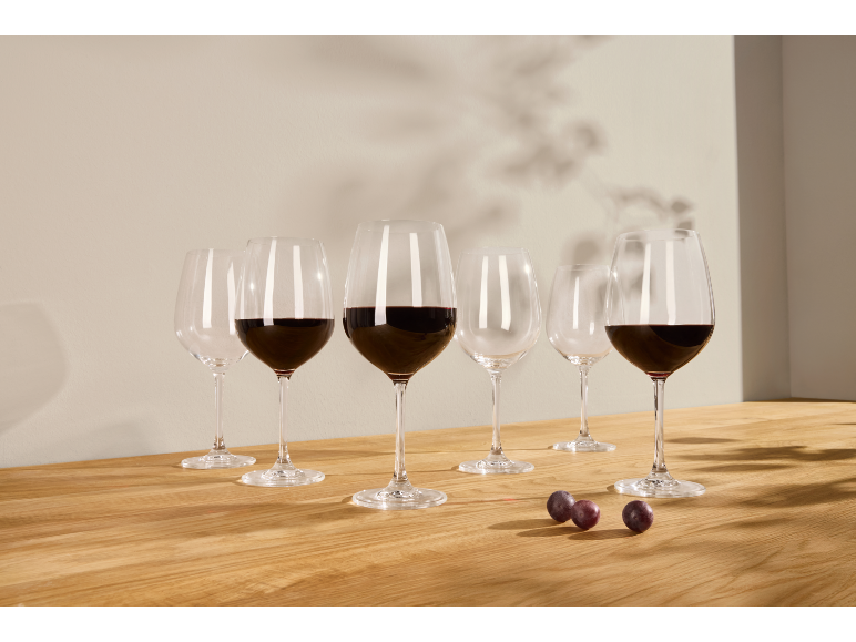 Verres de vin rouge, certains remplis, sur une table en bois avec des raisins et des ombres.