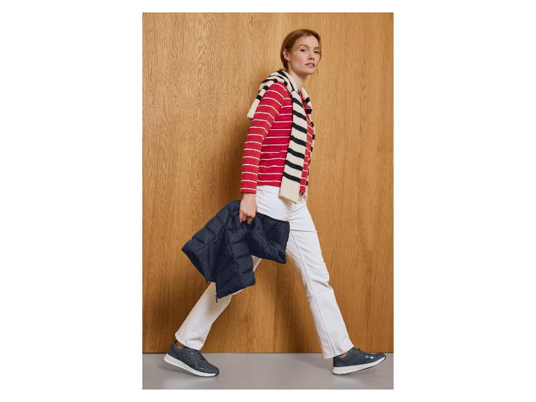 Femme en haut rayé rouge et blanc, pantalon blanc et baskets bleu foncé, tenant une veste bleu foncé.