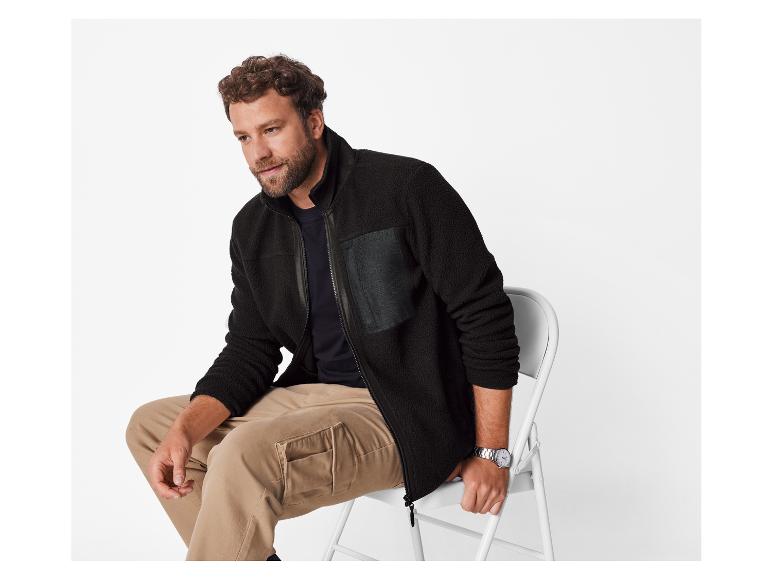 Un homme en veste polaire noire et pantalon kaki assis sur une chaise blanche.