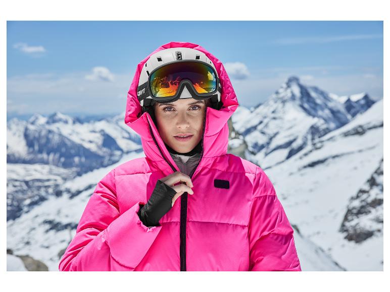 Femme en veste de ski rose et casque avec lunettes de ski sur fond de montagne enneigée.