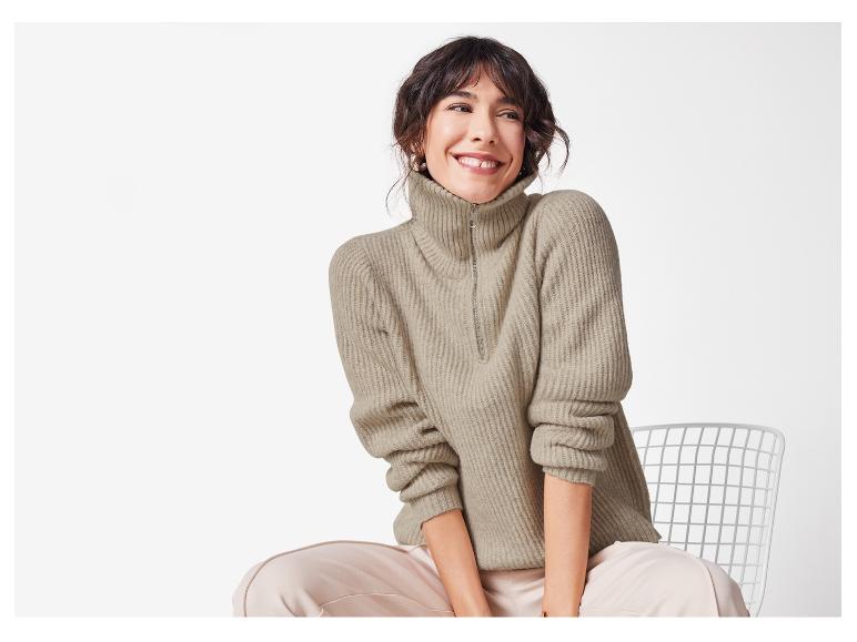 Une femme assise sur une chaise avec un pull beige à fermeture éclair et un pantalon rose clair.