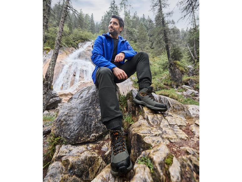 Homme en veste bleue et pantalon noir près d'une cascade, avec des chaussures de randonnée noires.