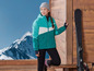 Une femme portant une veste de ski bleue regarde un ski appuyé contre un mur en bois.