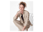 Modèle souriant assis sur une chaise, portant un ensemble pantalon et chemise beige.
