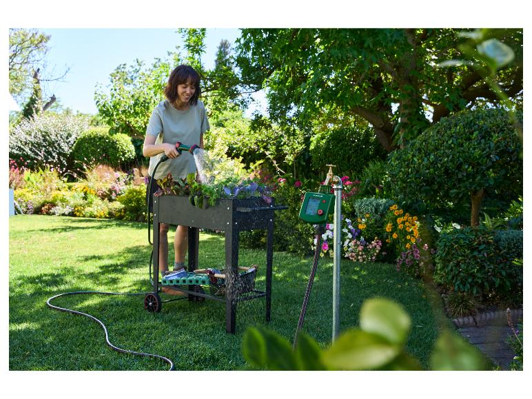Femme arrosant des plantes dans un potager surélevé avec un programmateur d'arrosage et un tuyau.