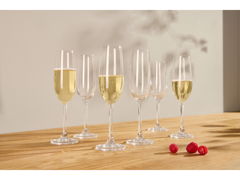 Six flûtes à champagne, certaines remplies de vin mousseux, et trois framboises sur une table en bois.