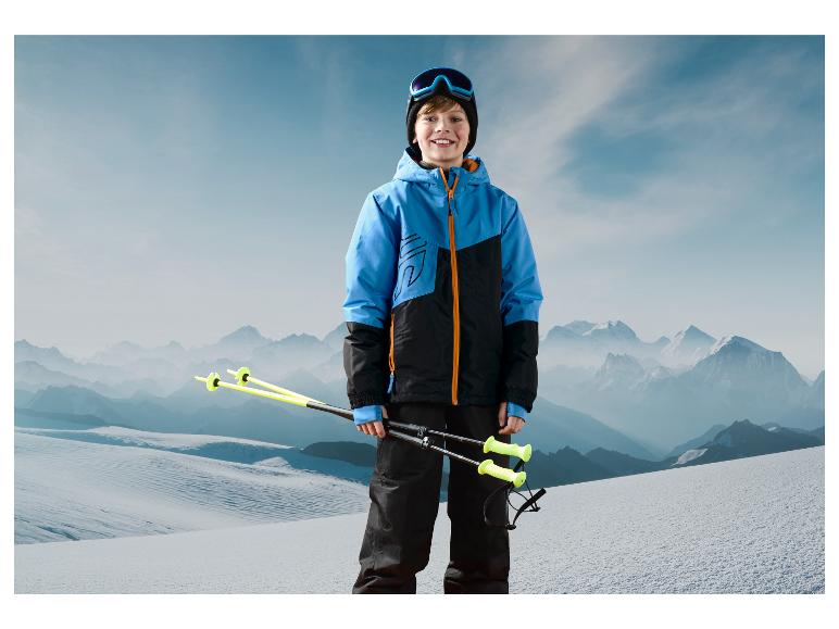 Un garçon en veste bleue et pantalon noir tient des bâtons de ski dans un paysage enneigé.