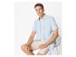 Homme en chemise bleu clair à manches courtes et short beige, assis sur une chaise.