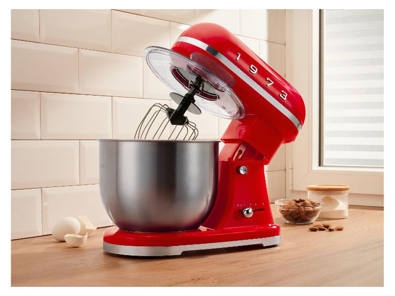 Robot pâtissier rouge avec bol, fouet, œufs et amandes sur un comptoir en bois.