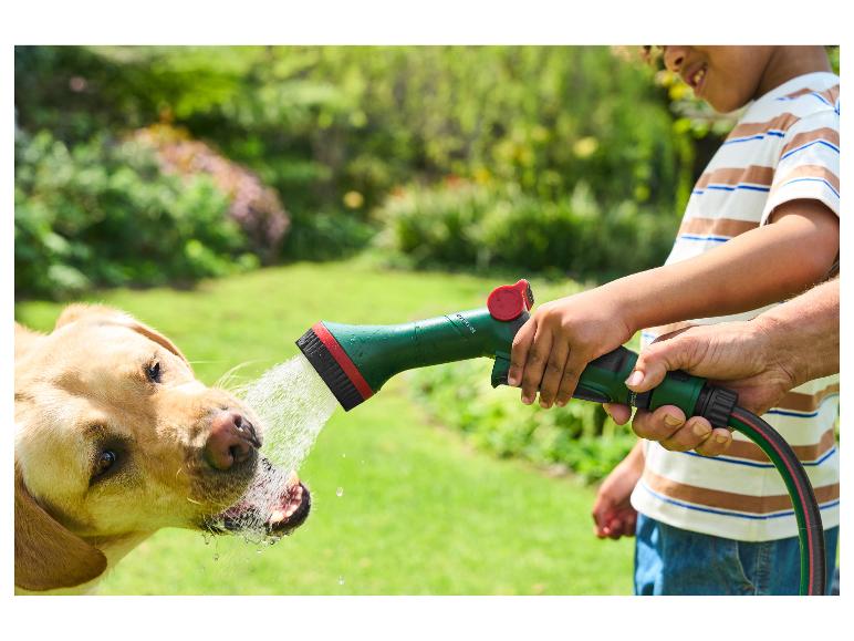 Enfant et adulte arrosant un chien avec un tuyau d'arrosage vert et un pistolet.