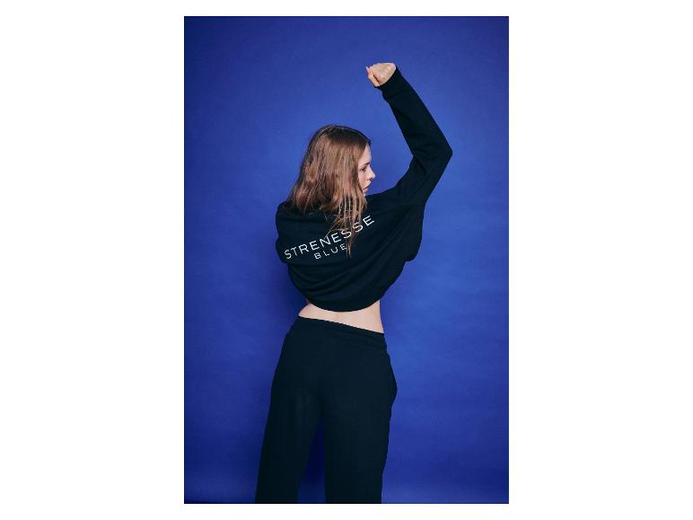 Femme en sweat à capuche et pantalon de survêtement noirs, bras levé, sur fond bleu.