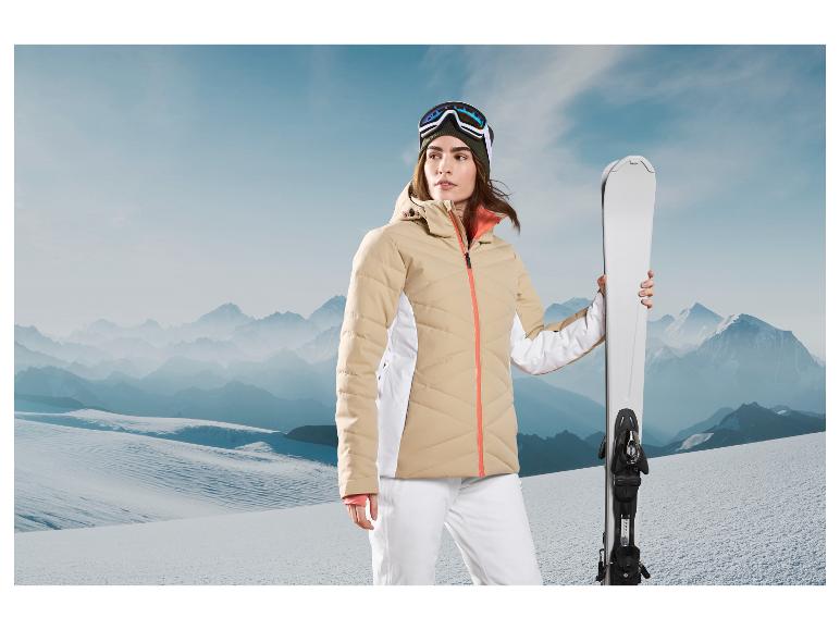 Une femme portant une veste de ski beige et un pantalon blanc, tenant des skis dans un paysage montagneux.