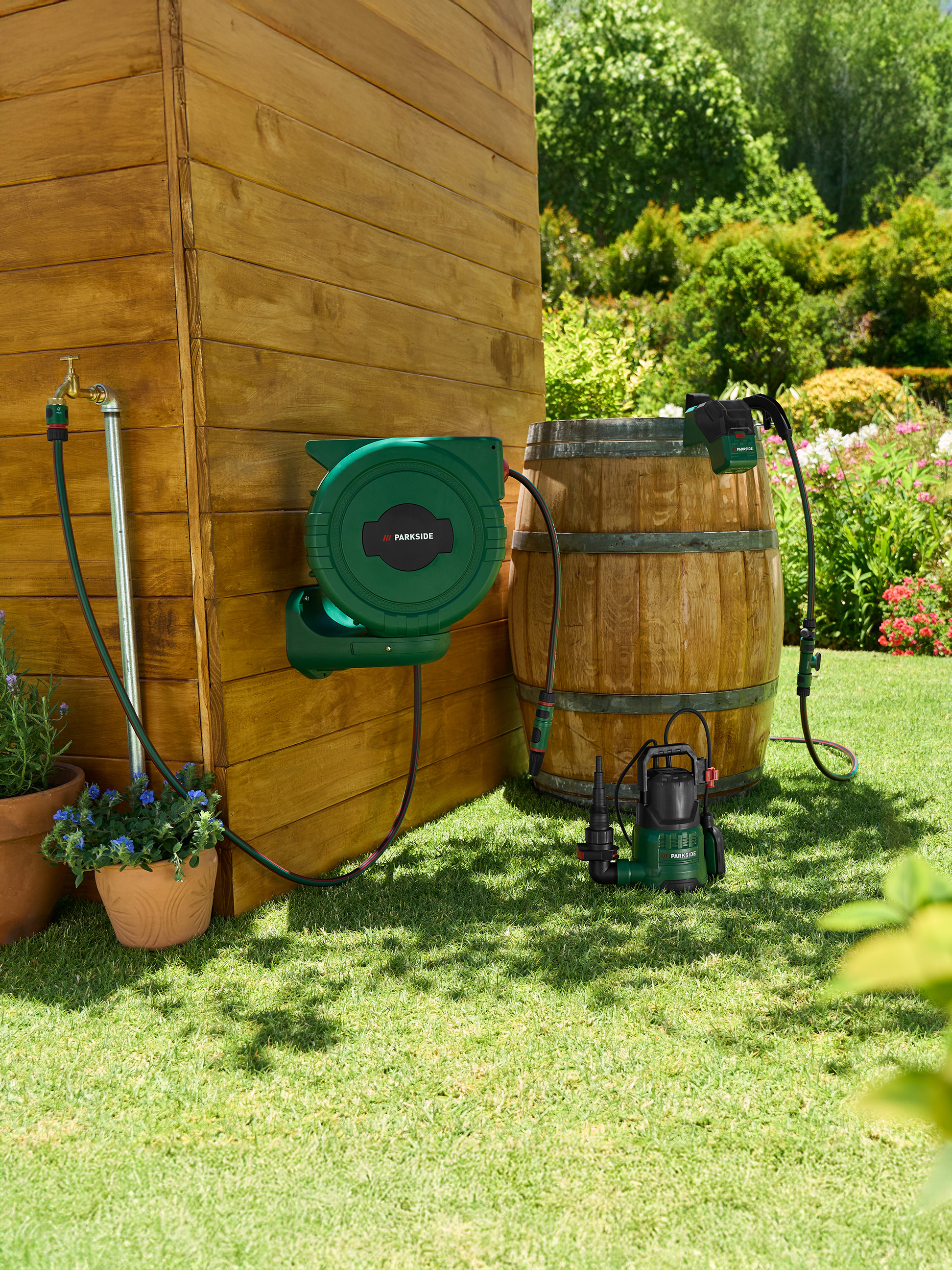 Dévidoir de tuyau d'arrosage et pompe près d'un abri en bois et d'un tonneau dans le jardin.