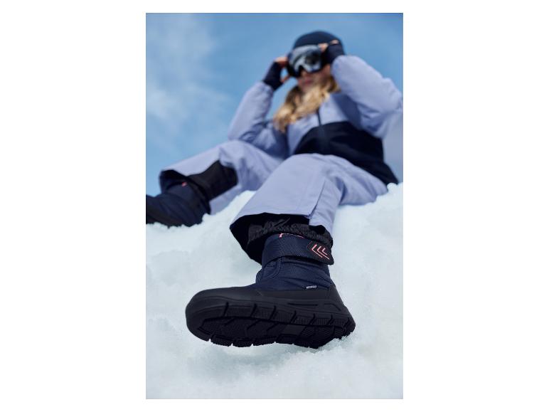 Une personne en pantalon de ski violet et bottes de neige bleu foncé assise dans la neige.