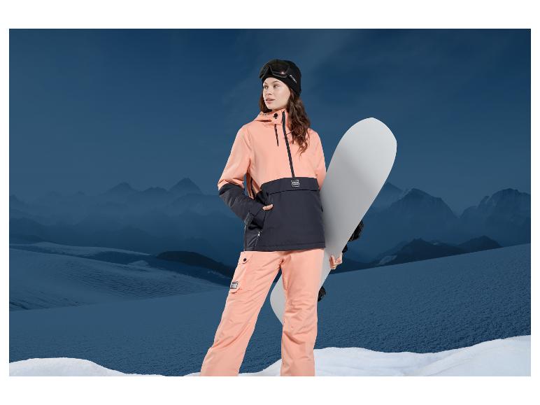 Femme en veste et pantalon de ski rose tenant un snowboard dans un paysage montagneux.
