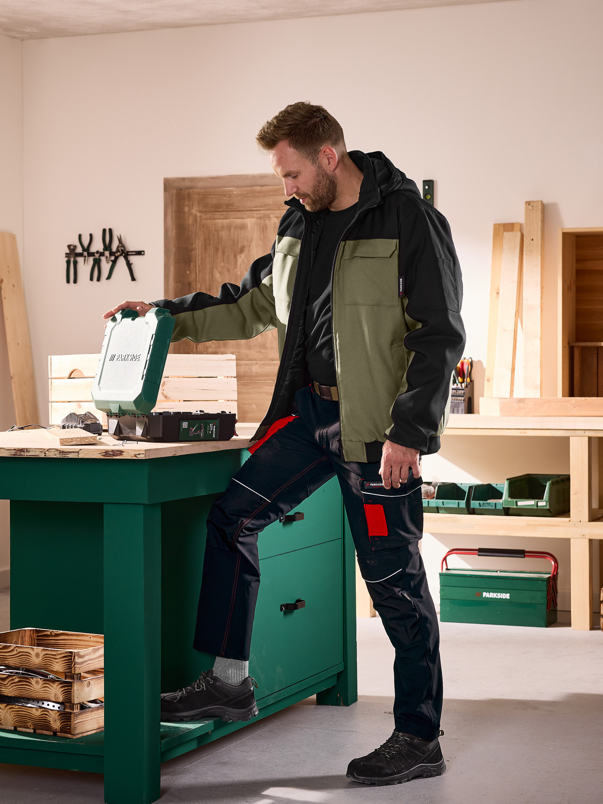 Homme en veste et pantalon de travail ouvrant une mallette à outils sur un établi dans un atelier.