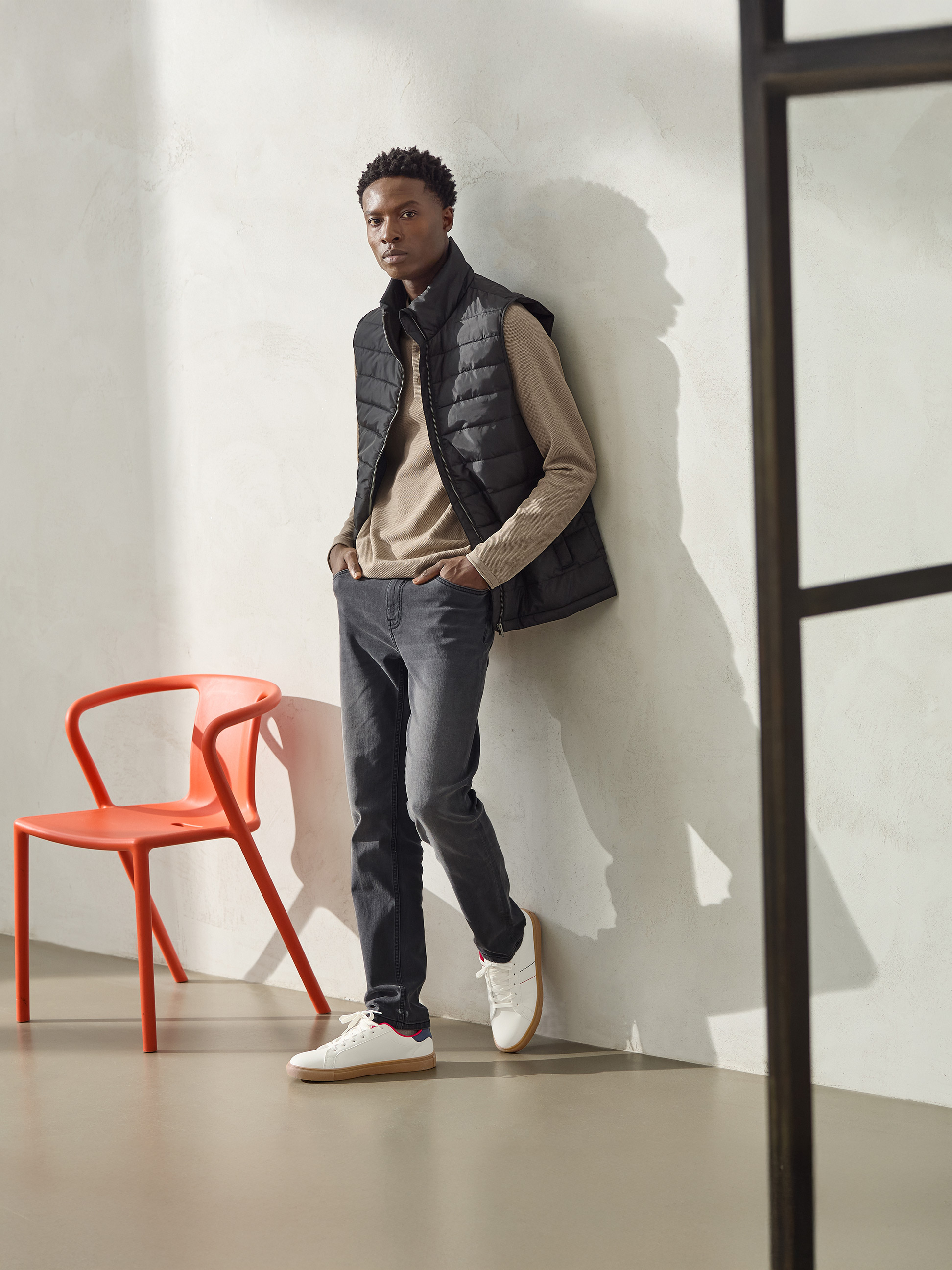 Homme en gilet matelassé noir, pull beige et jean gris, adossé à un mur.